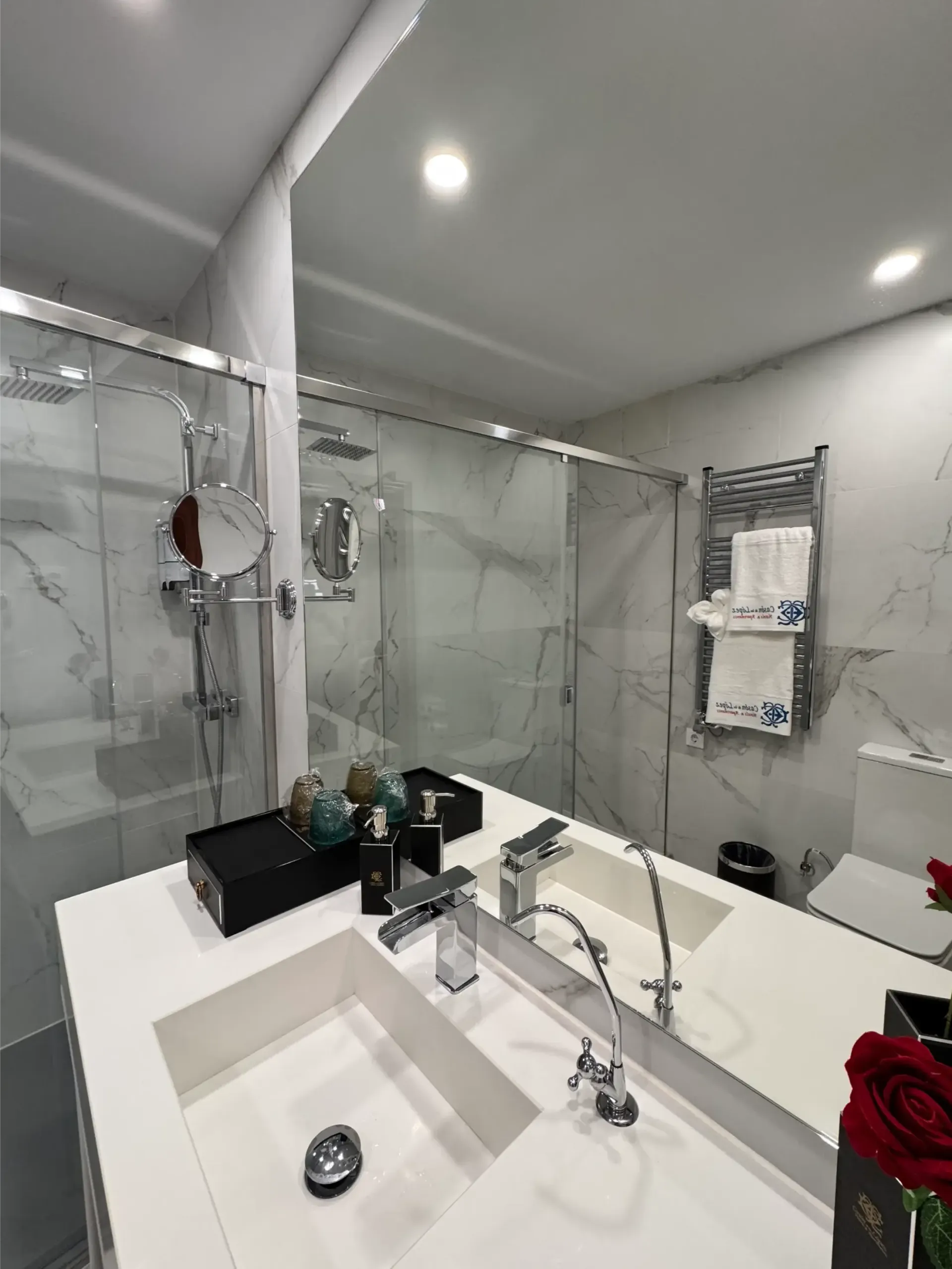 Baño moderno con lavabo doble, paredes con diseño de mármol, ducha de cristal y toallero eléctrico.