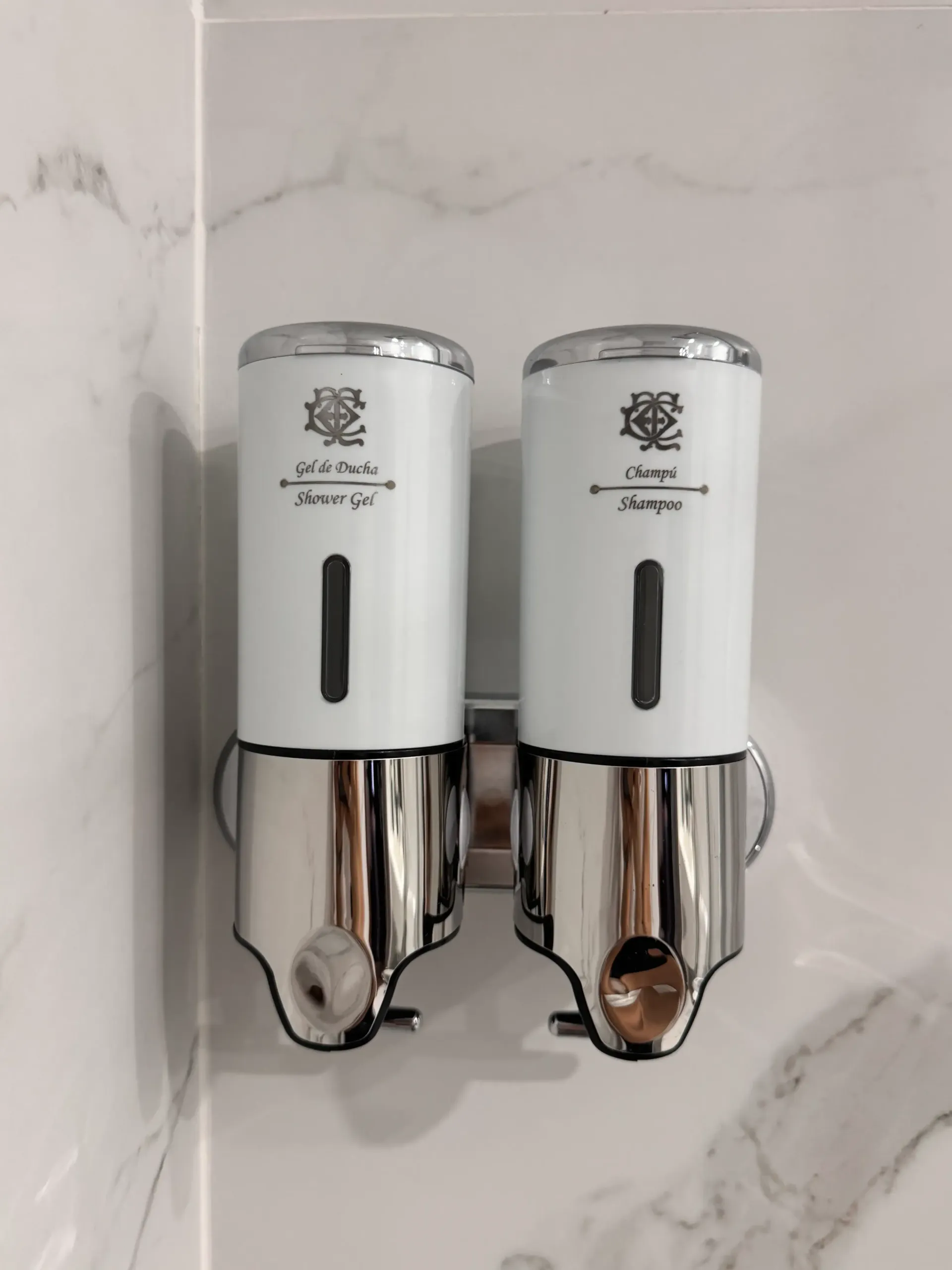 Un dispensador doble de jabón y champú para baño, montado en la pared, en color blanco y cromo, sobre una pared de azulejos con estampado de mármol blanco.