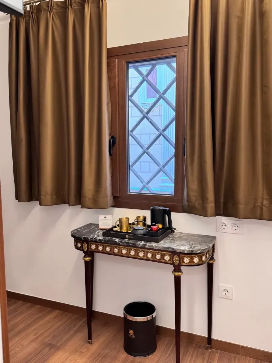 Una mesita auxiliar decorativa con un juego de té y una tetera debajo de una ventana con cortinas marrones en una habitación de hotel.