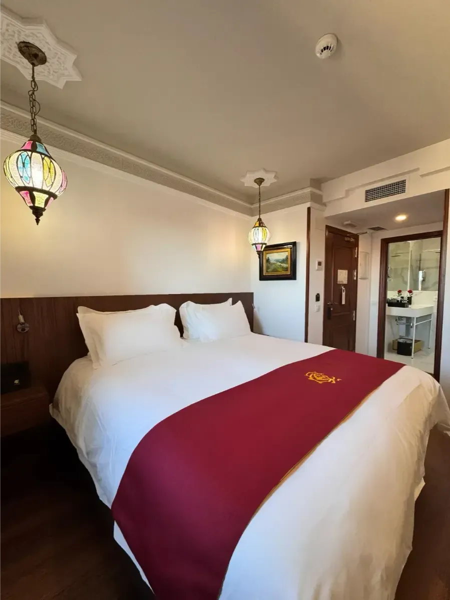 La habitación del hotel cuenta con una cama con sábanas blancas y un camino de mesa rojo, flanqueada por dos lámparas ornamentadas de vidrieras coloridas.