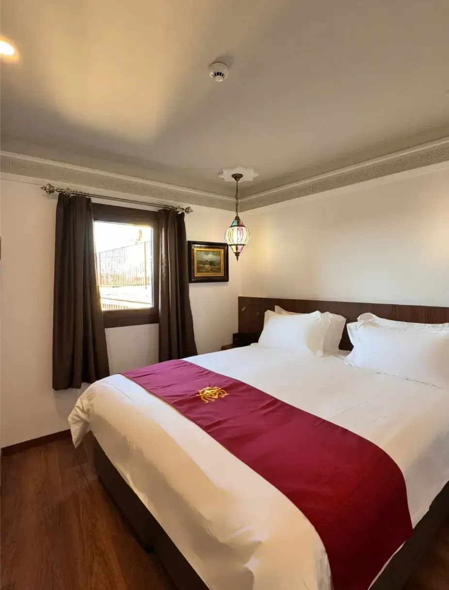 Una habitación de hotel con una cama grande, sábanas blancas, una alfombra color granate, cortinas oscuras y una lámpara colgante decorativa.