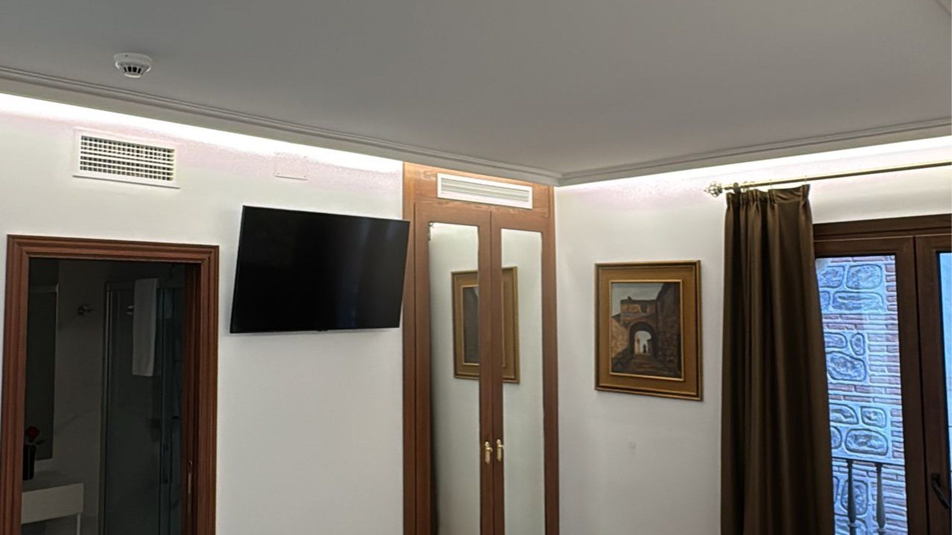 Un rincón de una habitación de hotel con un televisor montado en la pared, un armario con espejo, un cuadro enmarcado y una ventana con cortinas marrones.