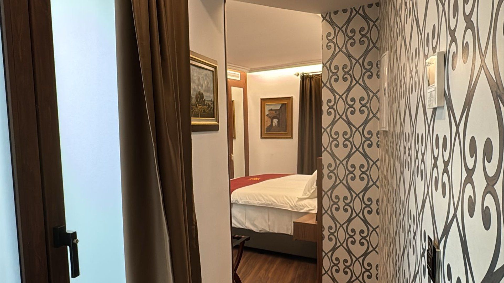 Vista a través de una puerta hacia un dormitorio con la cama hecha, cuadros enmarcados en la pared y papel pintado con estampado en el pasillo.