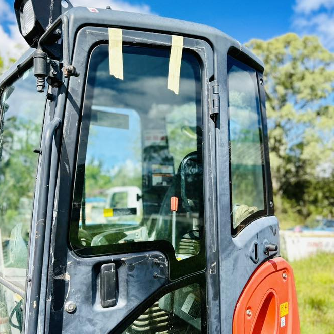 Close-up of a black excavator cab with a red machine arm in a sunny outdoor worksite.