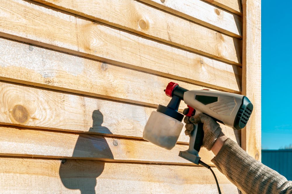 A Person Spray Painting Wooden Siding With An Electric Paint Sprayer — TMP Painting Services in Beaconsfield, QLD