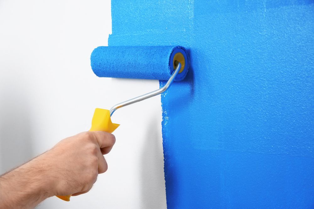 Hand using a paint roller to apply blue paint to a white wall — TMP Painting Services in Beaconsfield, QLD