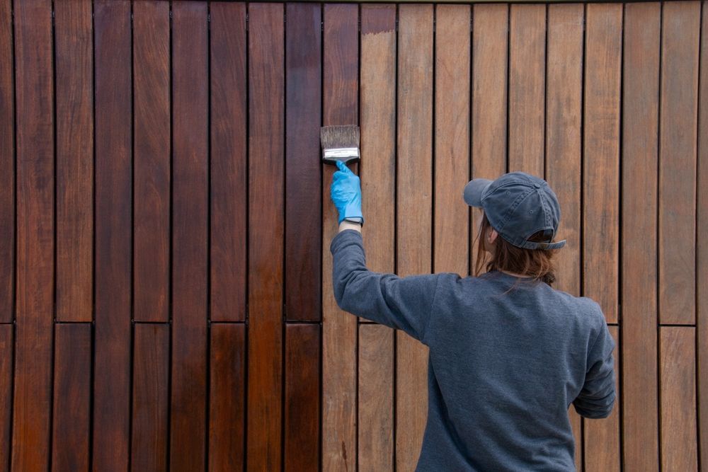 Person in a grey sweatshirt and cap staining a wooden fence with a brush, showing a color difference — TMP Painting Services in Beaconsfield, QLD