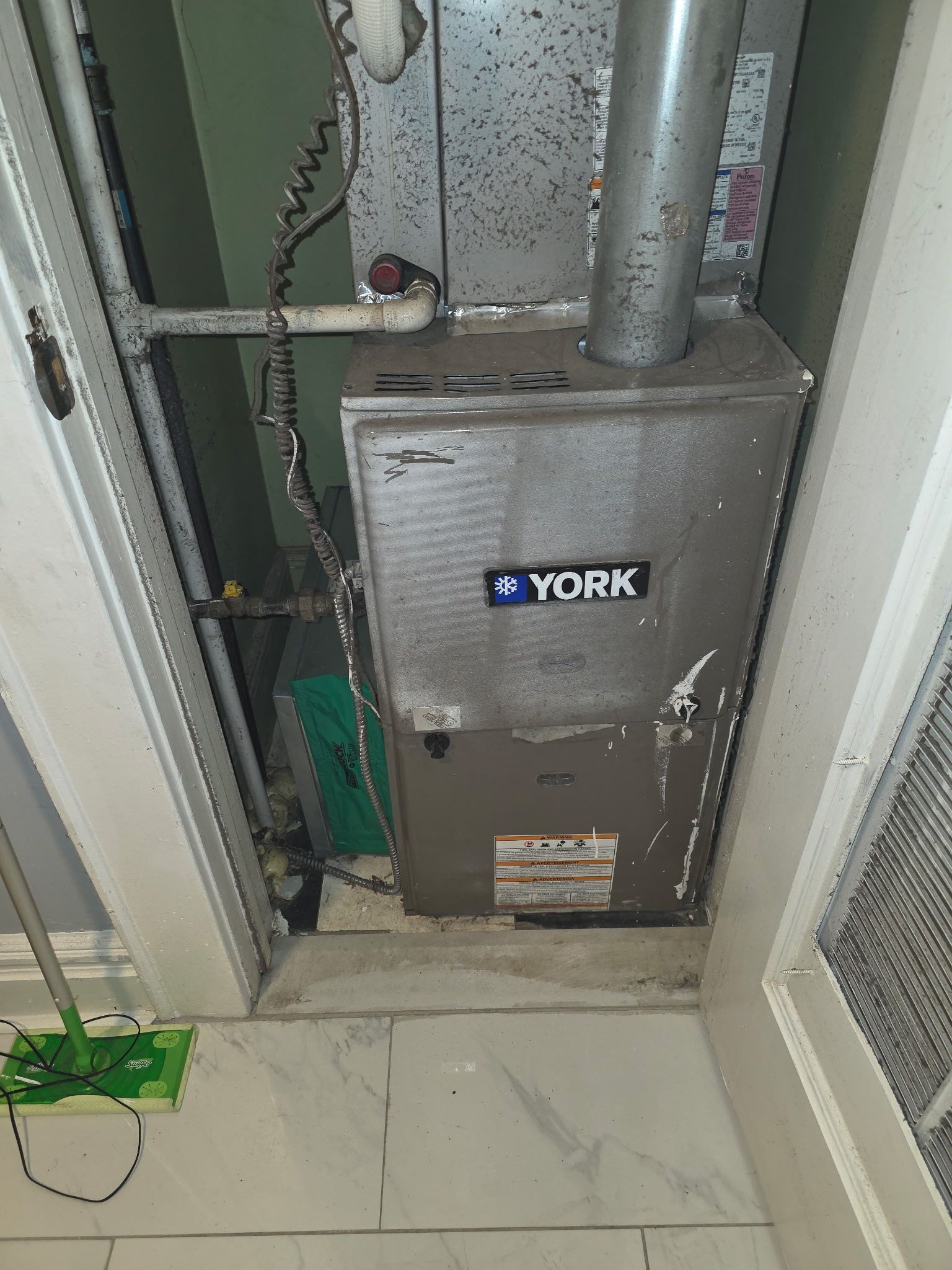 York furnace in a narrow utility closet with exposed pipes and tiled floor
