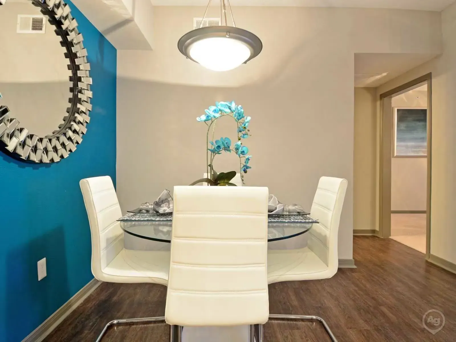 Dining area with round glass table and white upholstered chairs against a blue accent wall.