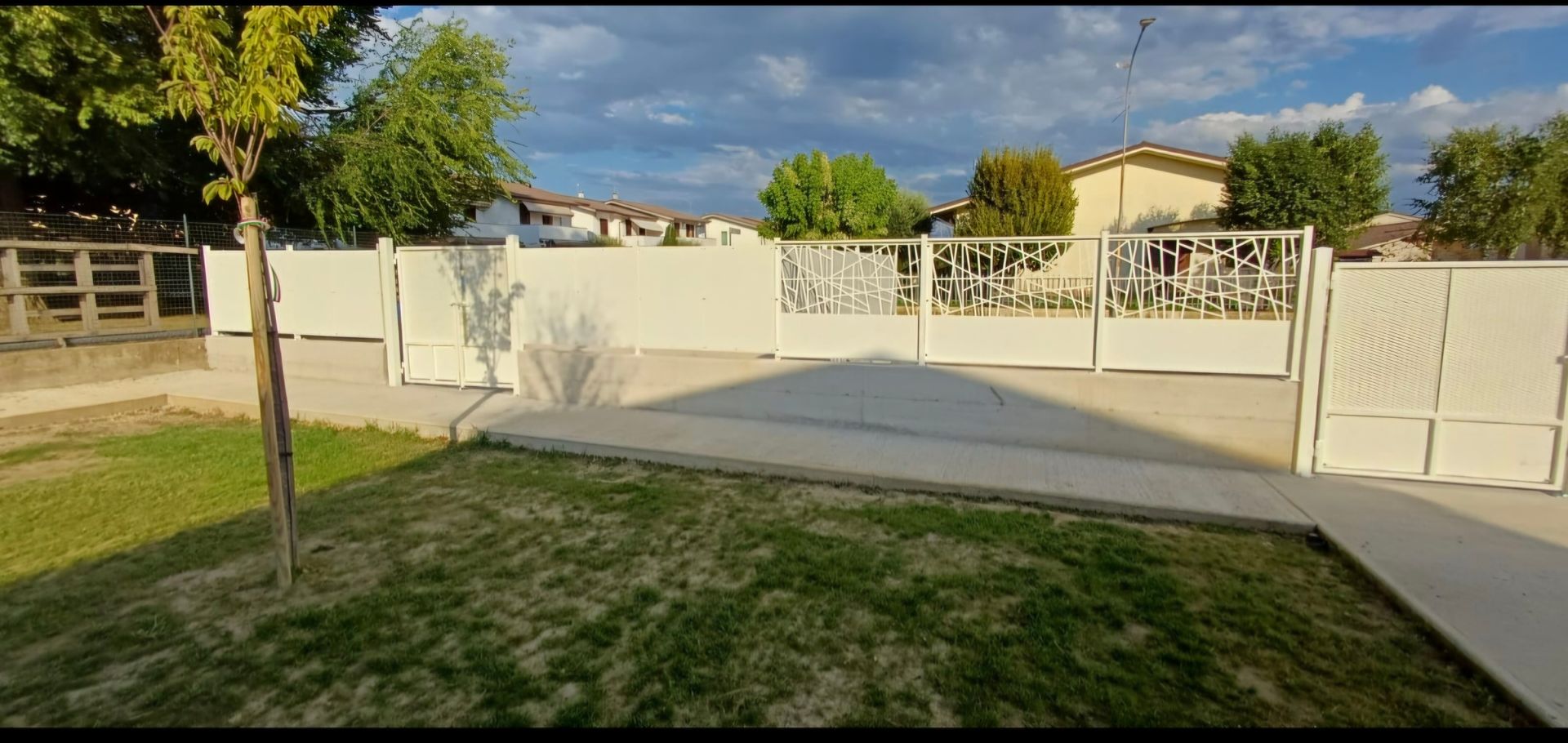 Una recinzione bianca davanti alle case con erba verde e un albero in primo piano.