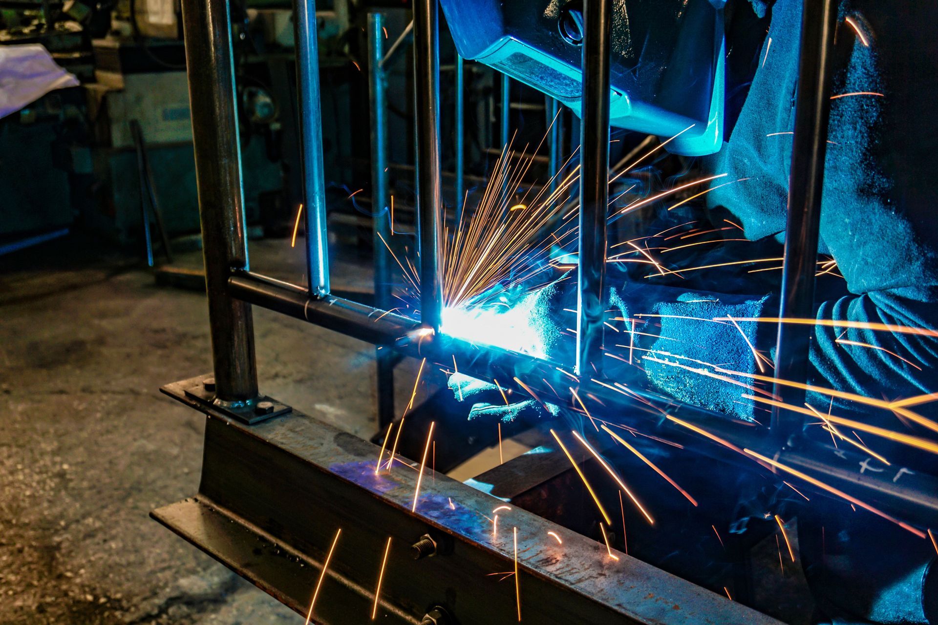 Un saldatore con indosso un equipaggiamento protettivo blu salda delle barre di metallo, creando scintille.
