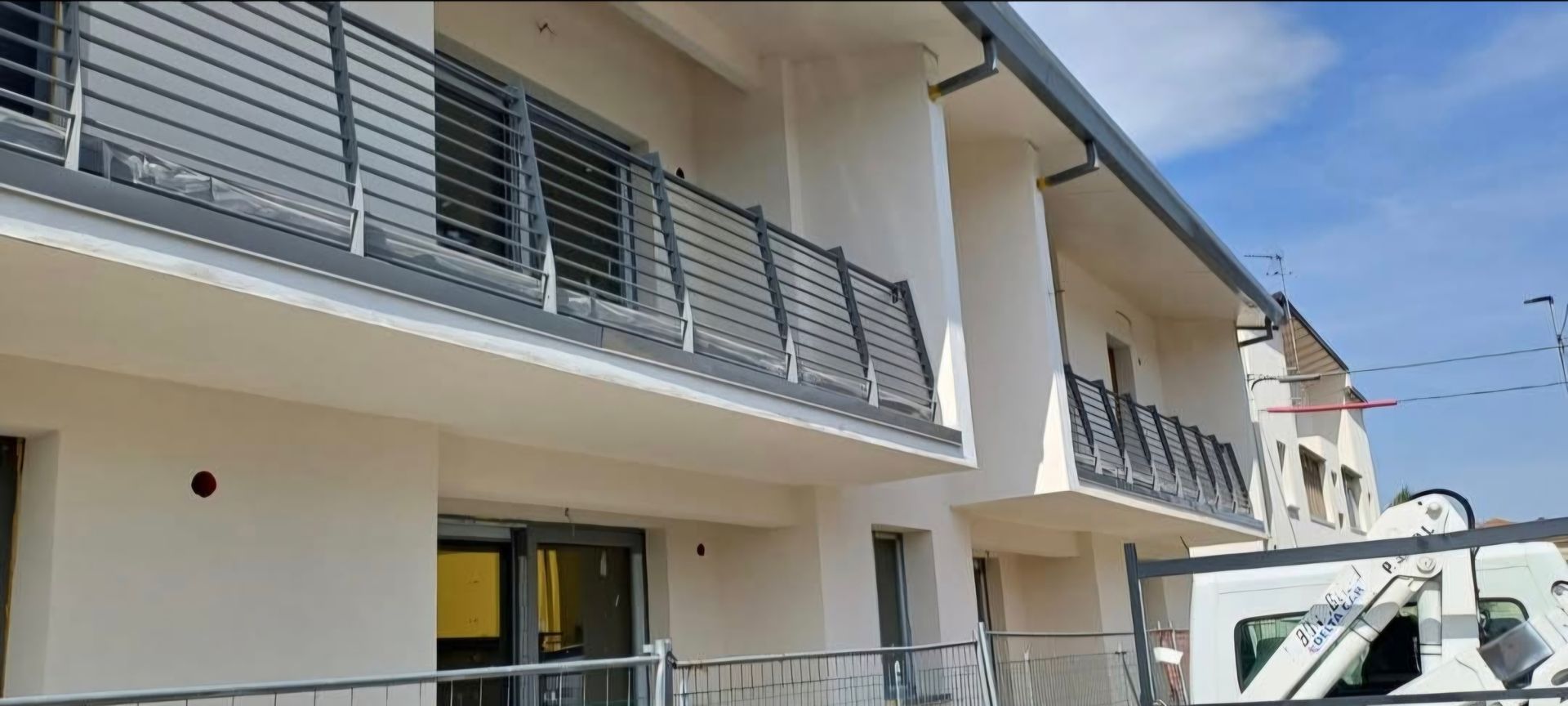 Edificio bianco a più piani con balconi e ringhiere metalliche sotto un cielo azzurro.