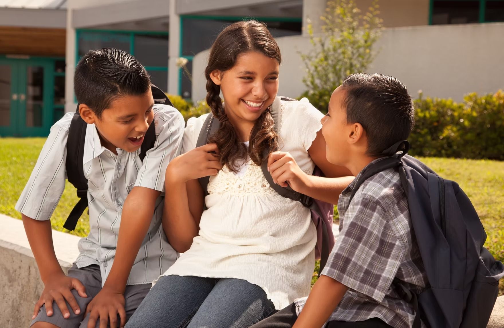 Three students with backpacks laugh together outside a school building on a sunny day.