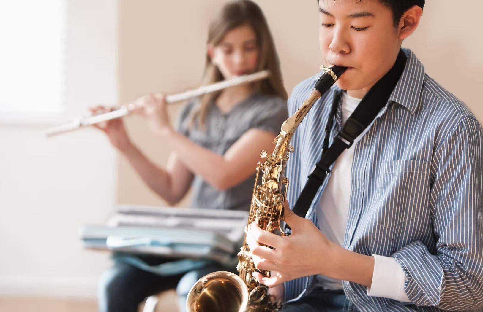 Boy playing saxophone, girl playing flute in a room. Both are looking down at their instruments while playing.