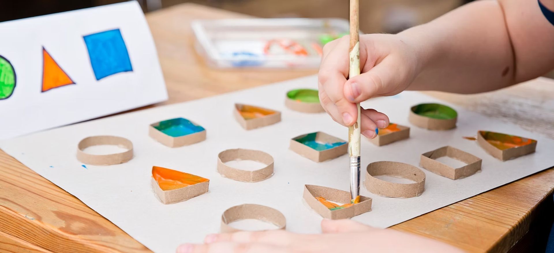 Child painting shapes with a brush. There are shape cutouts on a table, including circles, triangles, and squares.