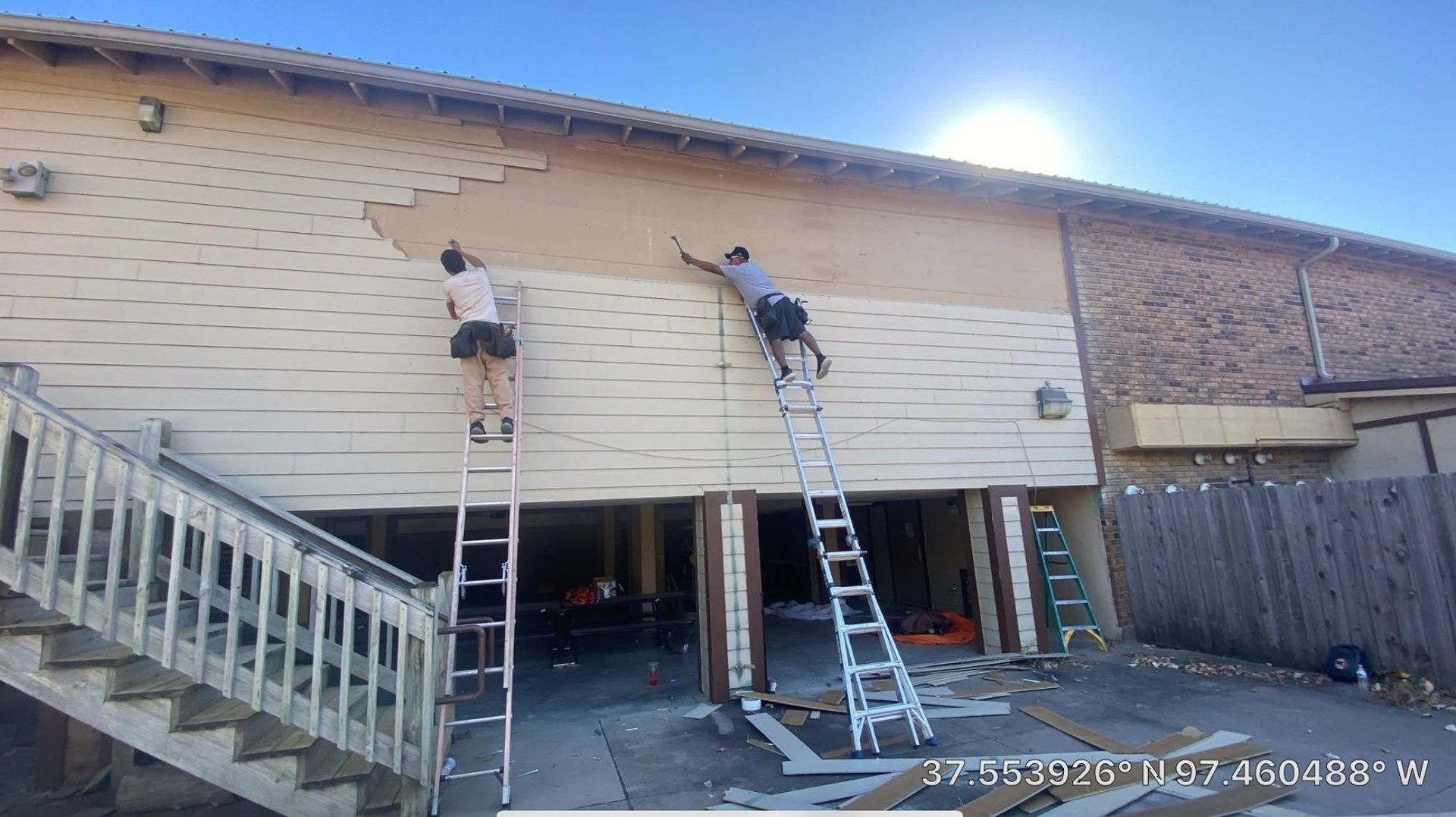 Two workers on ladders painting a beige building exterior above a driveway and stairway.