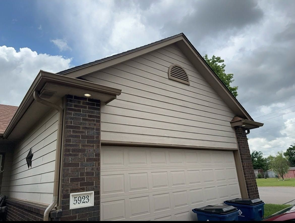 Suburban house exterior with beige siding, stone pillar, garage door, and blue trash bins by the driveway