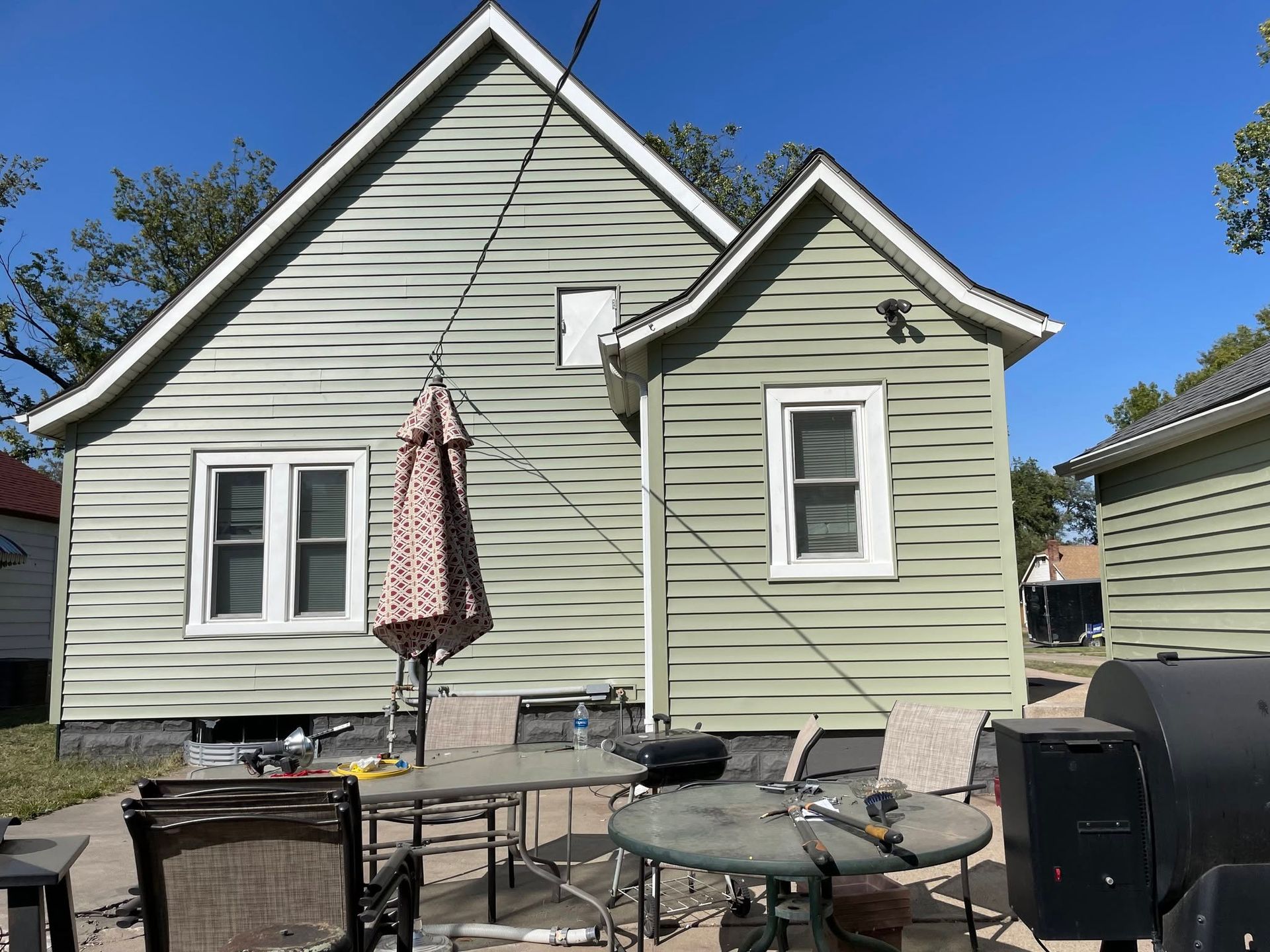 Backyard patio with glass table, umbrella, grill, and pale green house siding under a blue sky