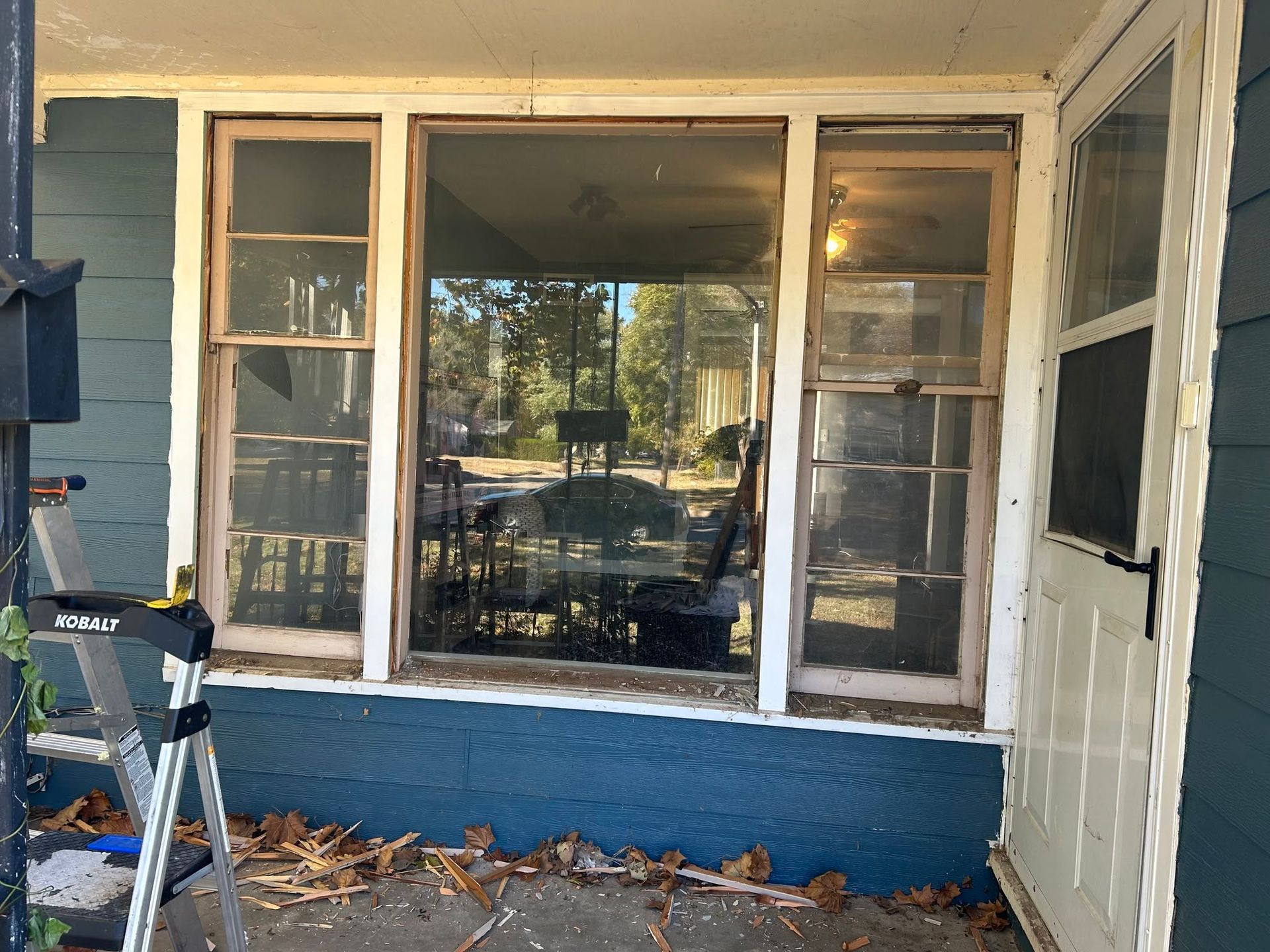 Front porch with a large window, blue lower wall, and a step ladder beside dried leaves.