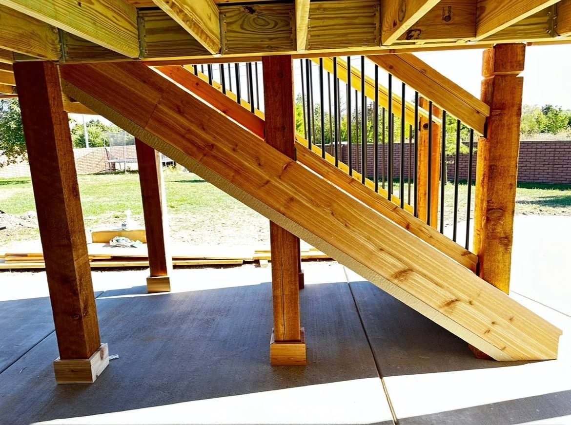 Wooden staircase and support posts under a raised deck, with sunlight and a concrete patio below