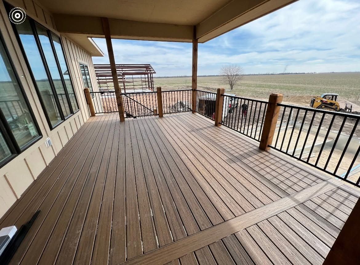 Covered wooden deck with black railing overlooking open fields and a cloudy horizon