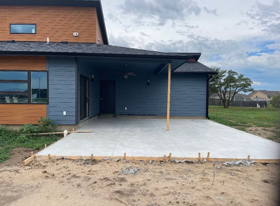 Modern house exterior with blue siding, wood accents, and a covered concrete driveway under construction
