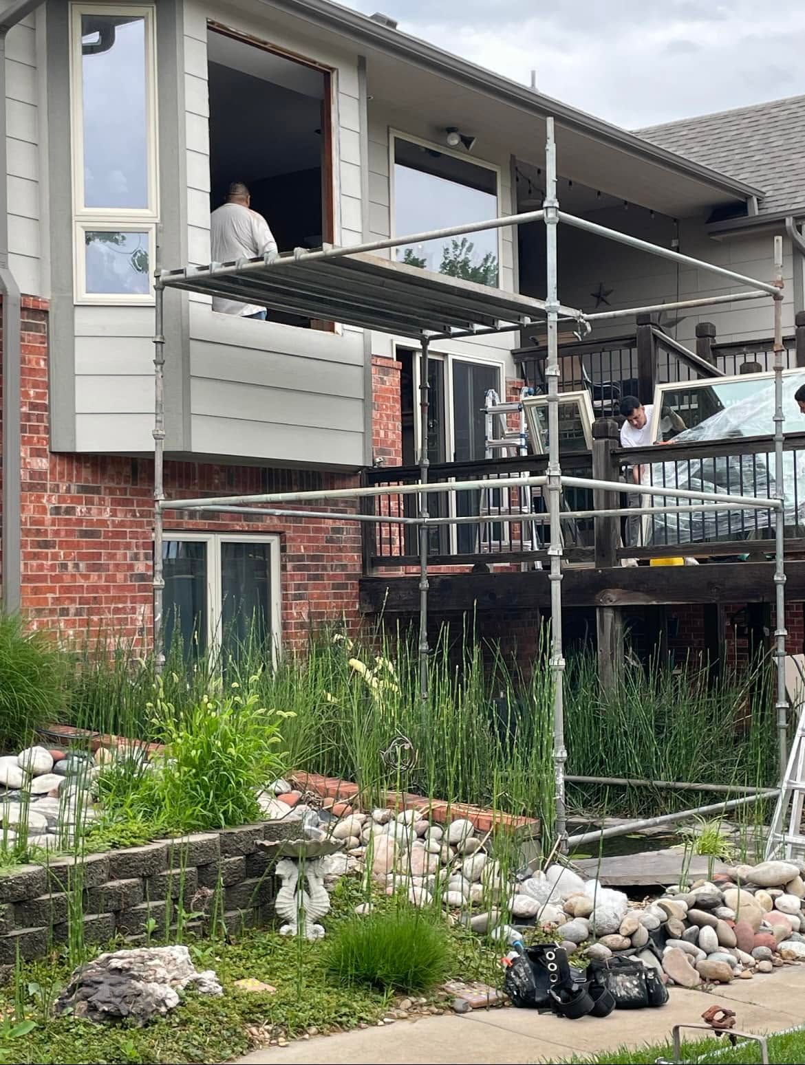 Scaffolding beside a house during exterior renovation, with weeds and rocks in the foreground