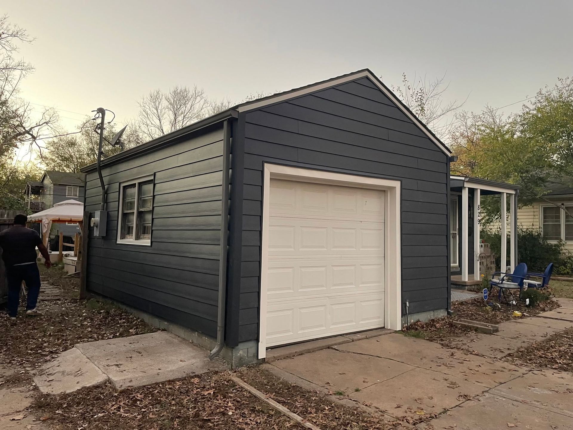 Dark gray detached garage with white door, small windows, and bare yard at dusk