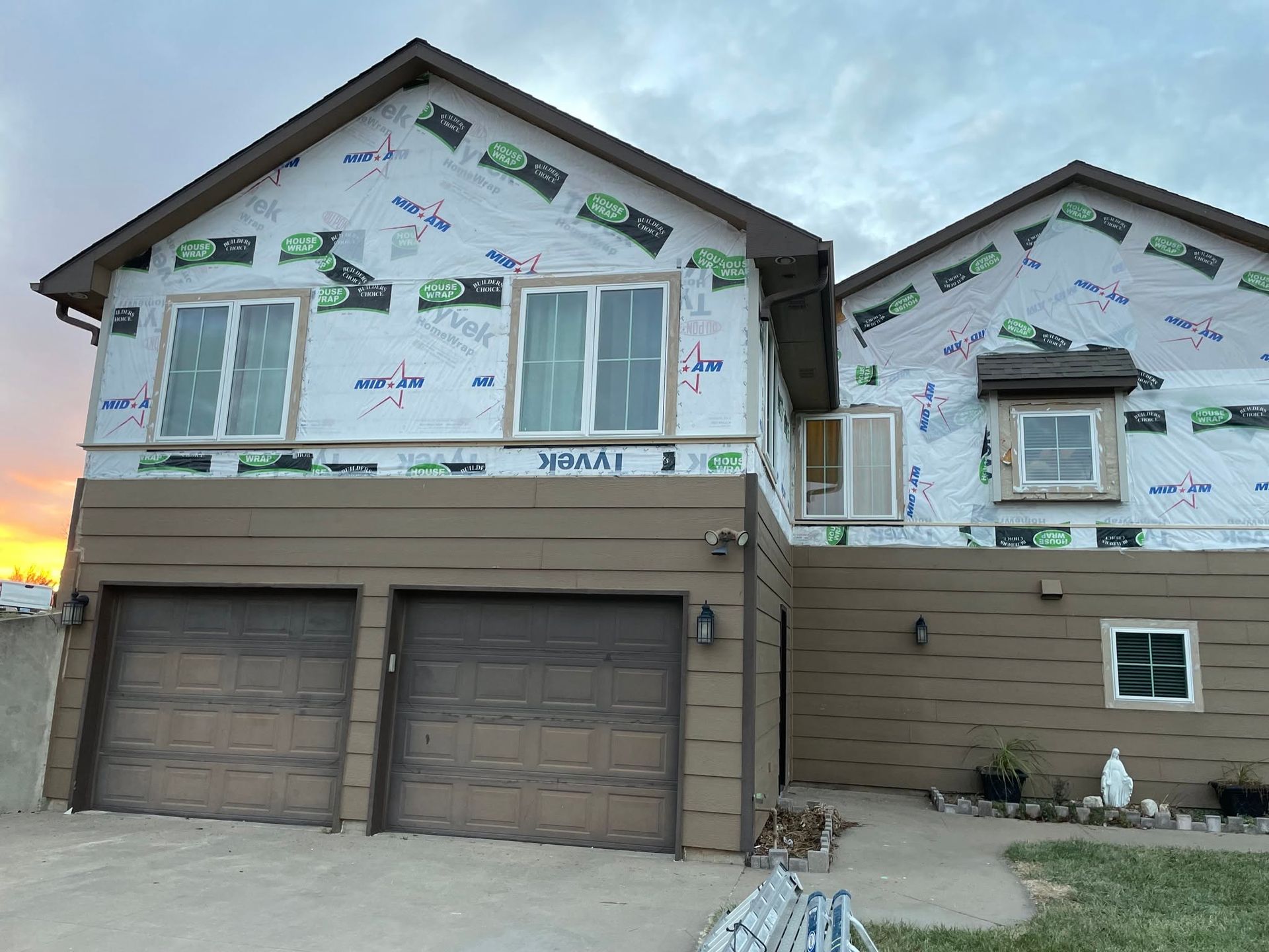 Two-story house with garage under construction, wrapped in white housewrap and tan siding at sunset