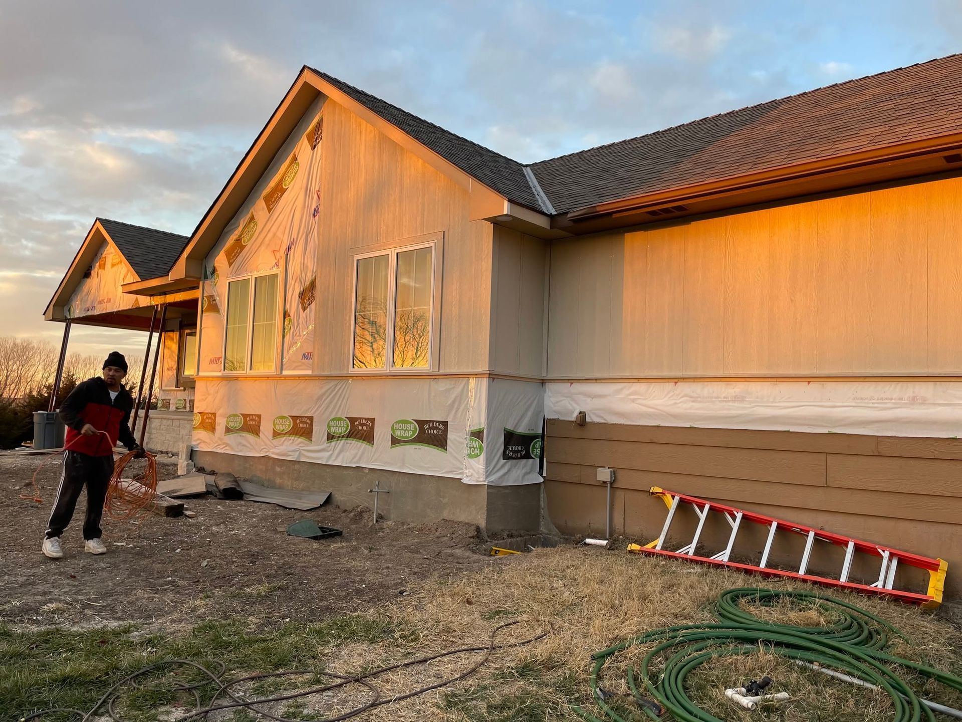Person painting the exterior of a house at sunset, with ladders and tools along the wall.