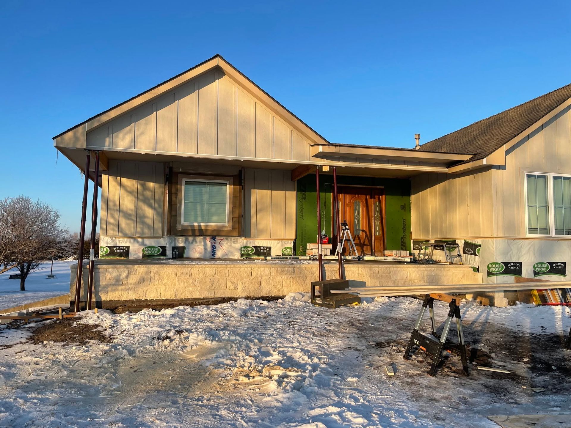 Snowy front yard with a beige house, porch, and green door under a blue sky