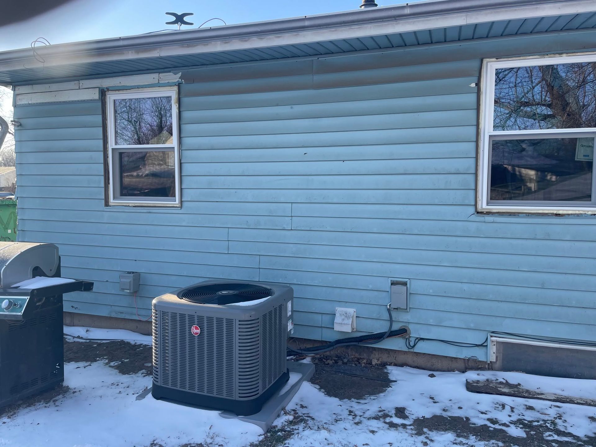 Blue house exterior with snow on the ground and an HVAC unit beneath two windows.
