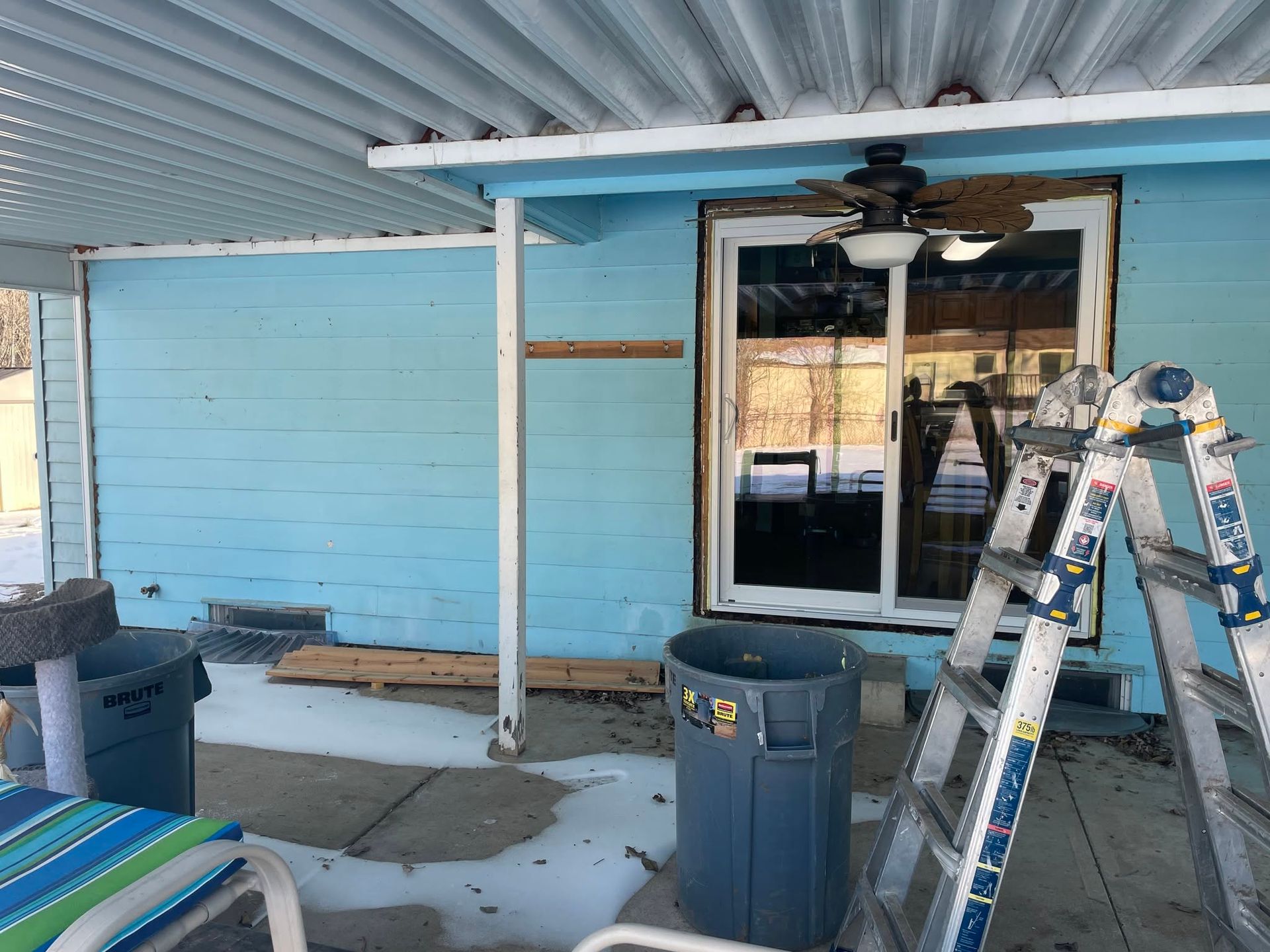Blue house porch with a sliding glass door, ceiling fan, trash can, and a ladder under a corrugated roof.
