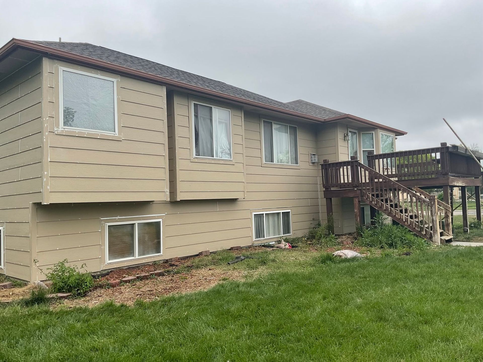 Beige split-level house with brown deck and stairs, viewed from a grassy yard under overcast sky.