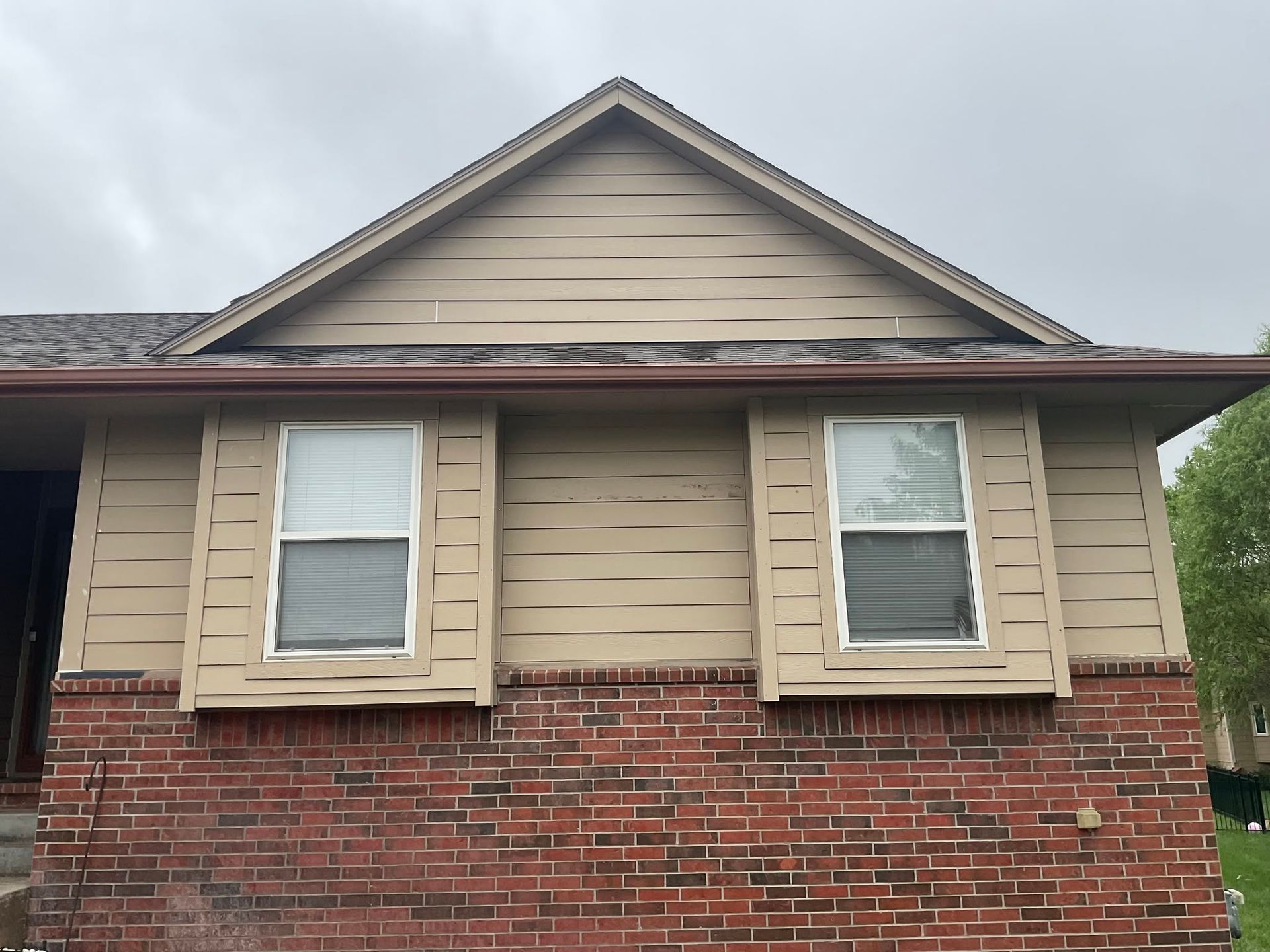 Tan house with red brick lower facade and two front windows under a cloudy sky