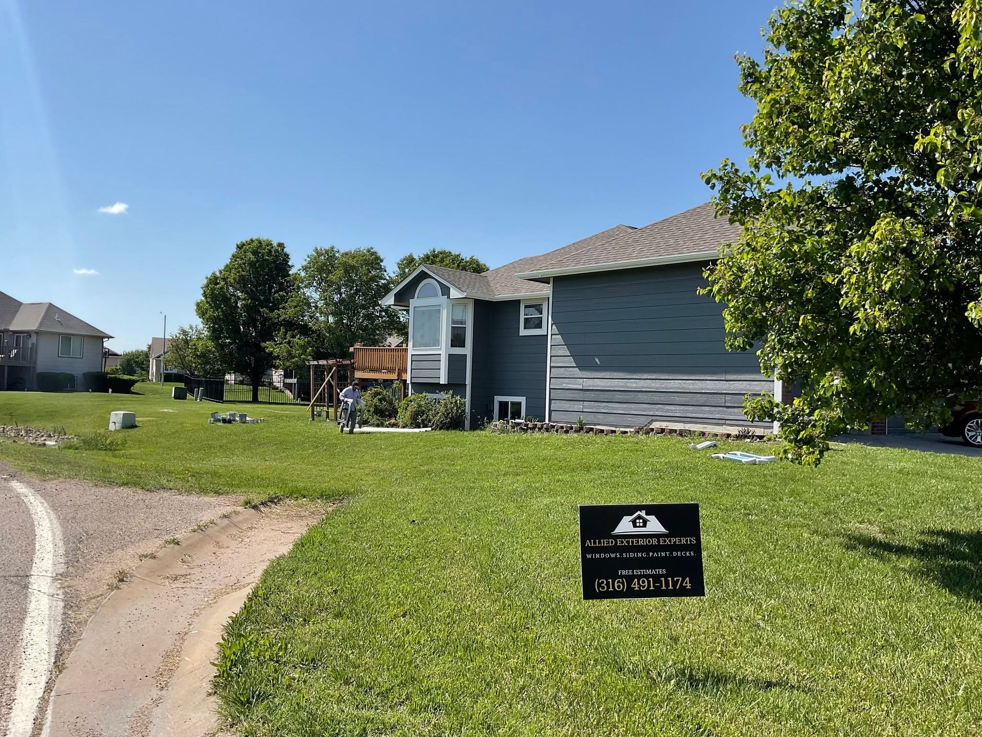 Suburban house with blue siding, green lawn, and a real estate sign by the curb under a clear sky.