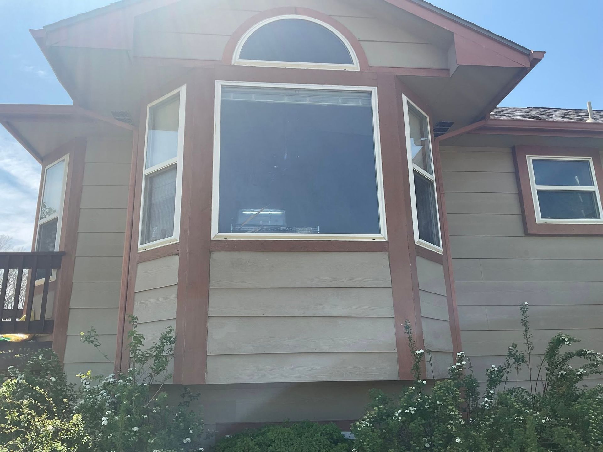 Front exterior of a house with brown siding, a large bay window, and red trim under bright sunlight