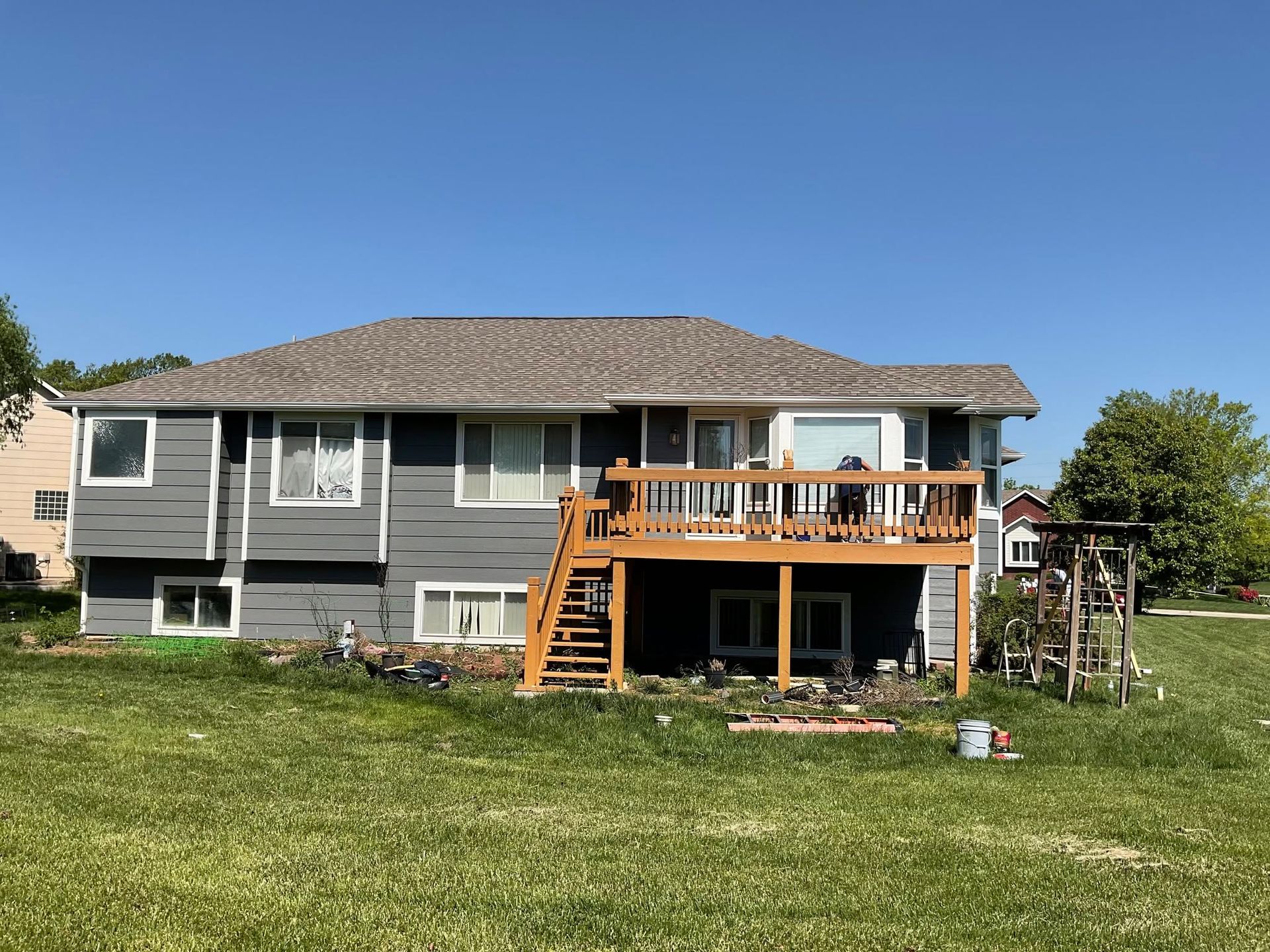 Gray house with a wooden deck and stairs in a grassy yard under a blue sky