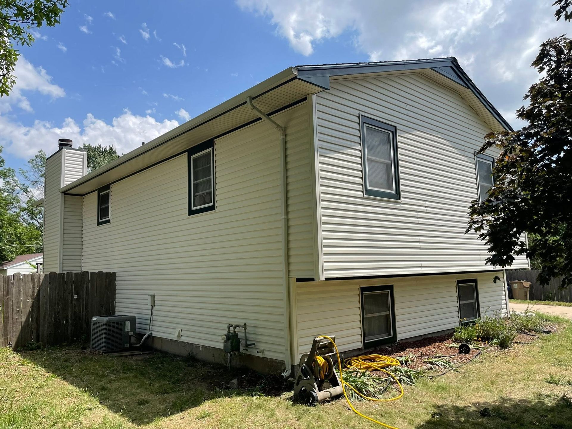 Side view of a beige house with white trim, small basement windows, and a grassy yard under a blue sky.