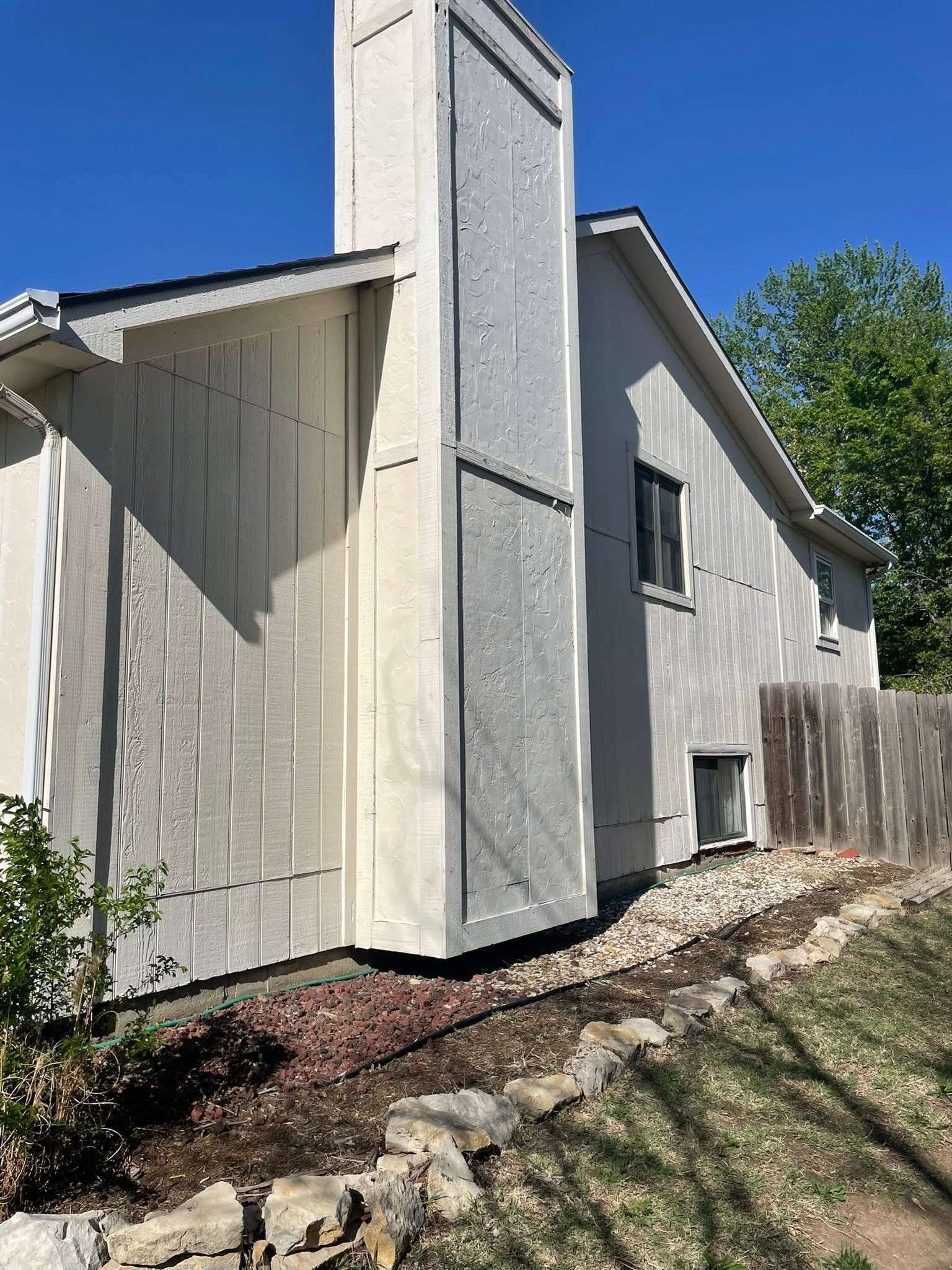 Side view of a gray house with a tall chimney, white trim, and a rock border along the foundation.