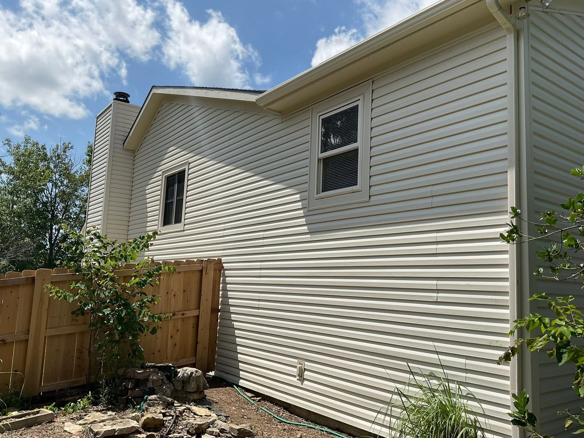 Side of a white vinyl-sided house with small windows, wooden fence, and a sunny backyard with trees and blue sky.