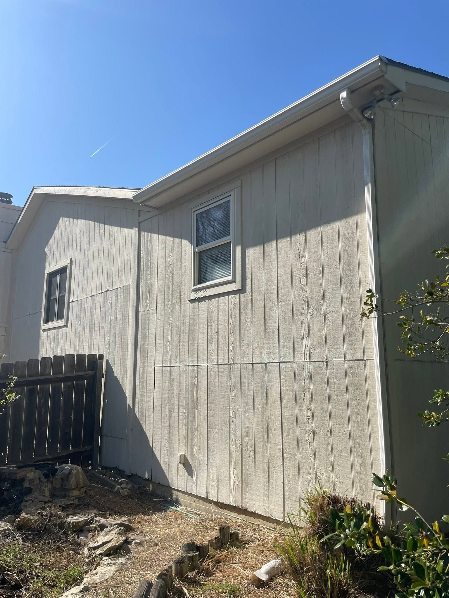Exterior of a light-colored house with two windows, a fence, and shrubs under a clear blue sky
