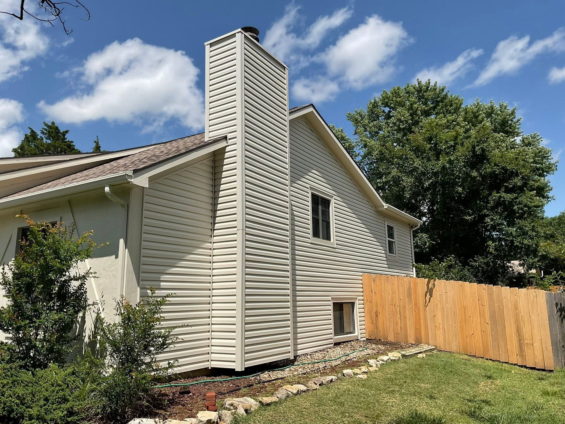 Side view of a beige house with a stone chimney, fenced yard, green lawn, and blue sky with clouds