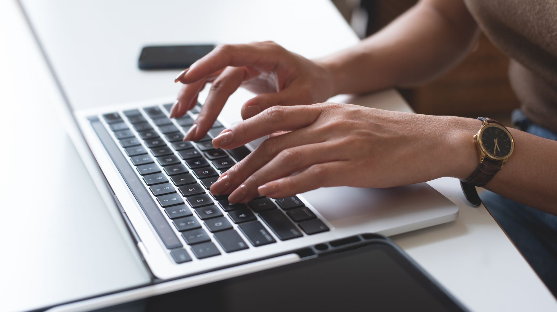 Hands typing on a silver laptop keyboard at a desk, with a watch on one wrist and a tablet nearby.