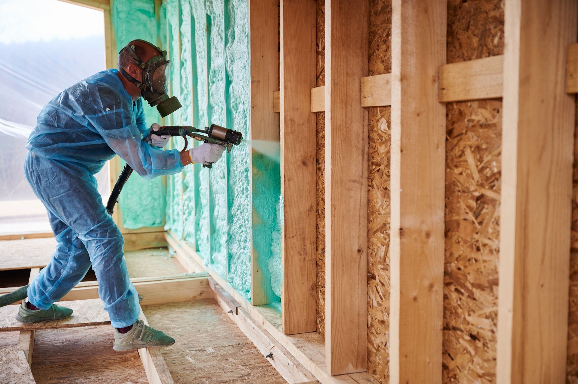 Person in protective gear sprays foam insulation onto a wooden wall in a building.