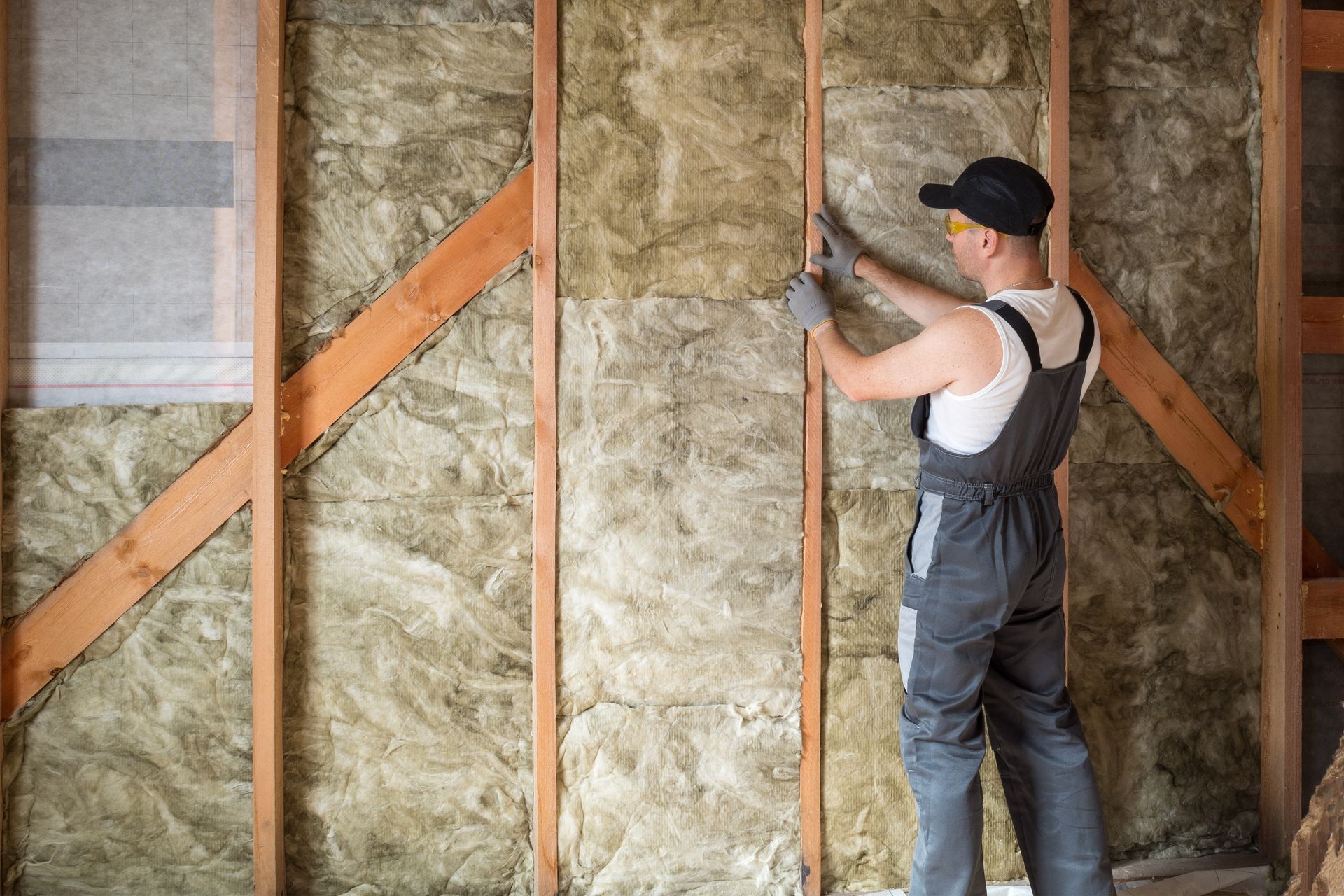 Man installing insulation between wooden wall studs, wearing safety gear.