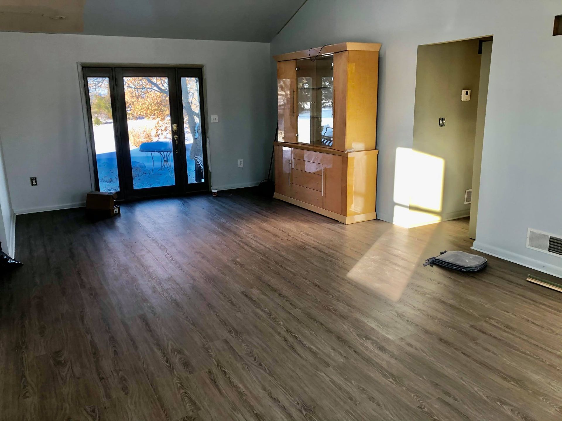 An empty room with gray wood-look flooring, light walls, a wooden china cabinet, and a black-framed sliding glass door.