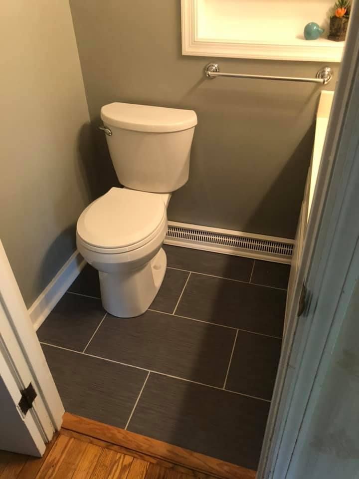 A white toilet in a small bathroom with grey walls, dark rectangular floor tiles, and a white baseboard.