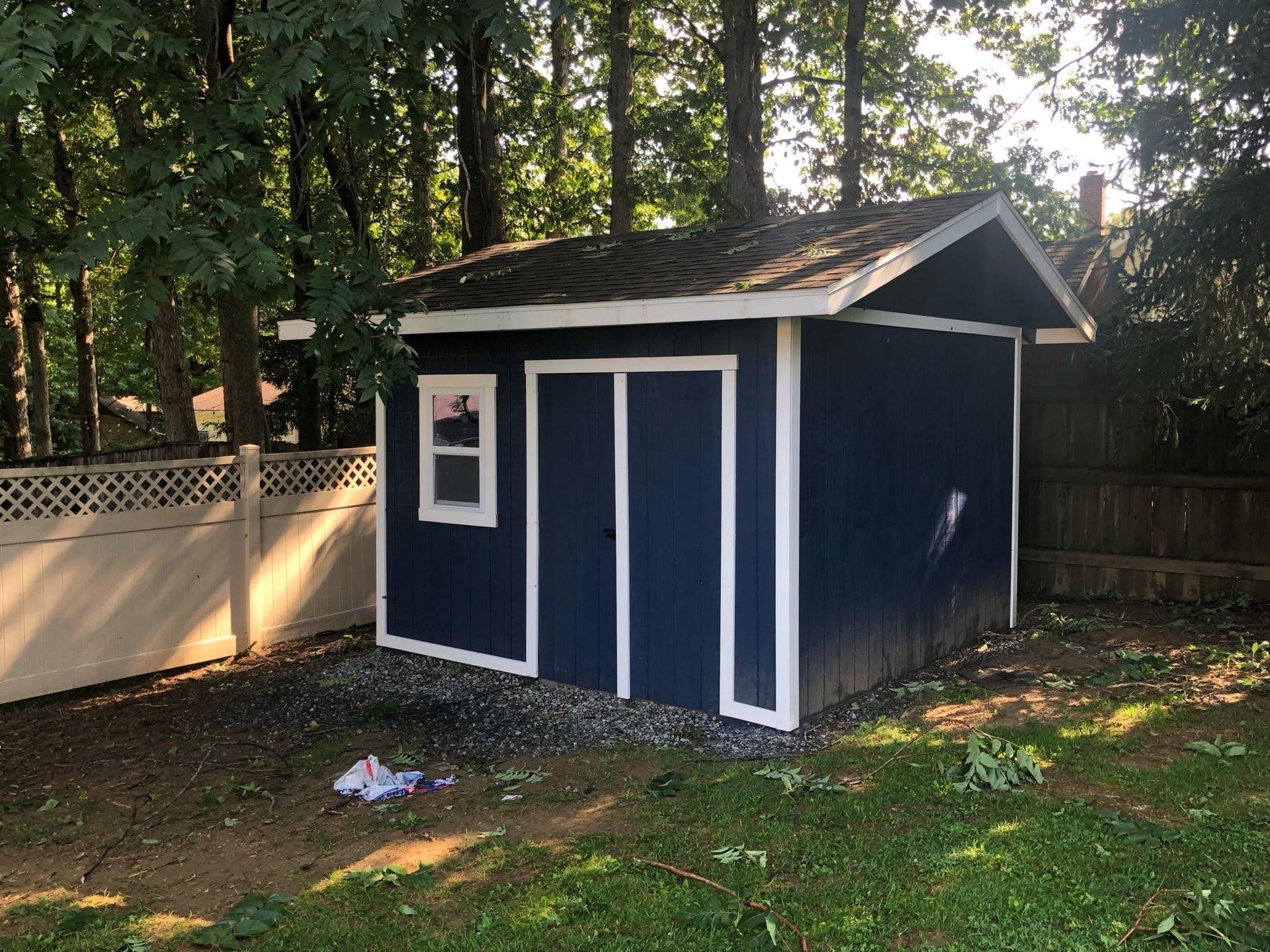 A dark blue storage shed with white trim and a single window sits in a backyard next to a lattice fence and trees.
