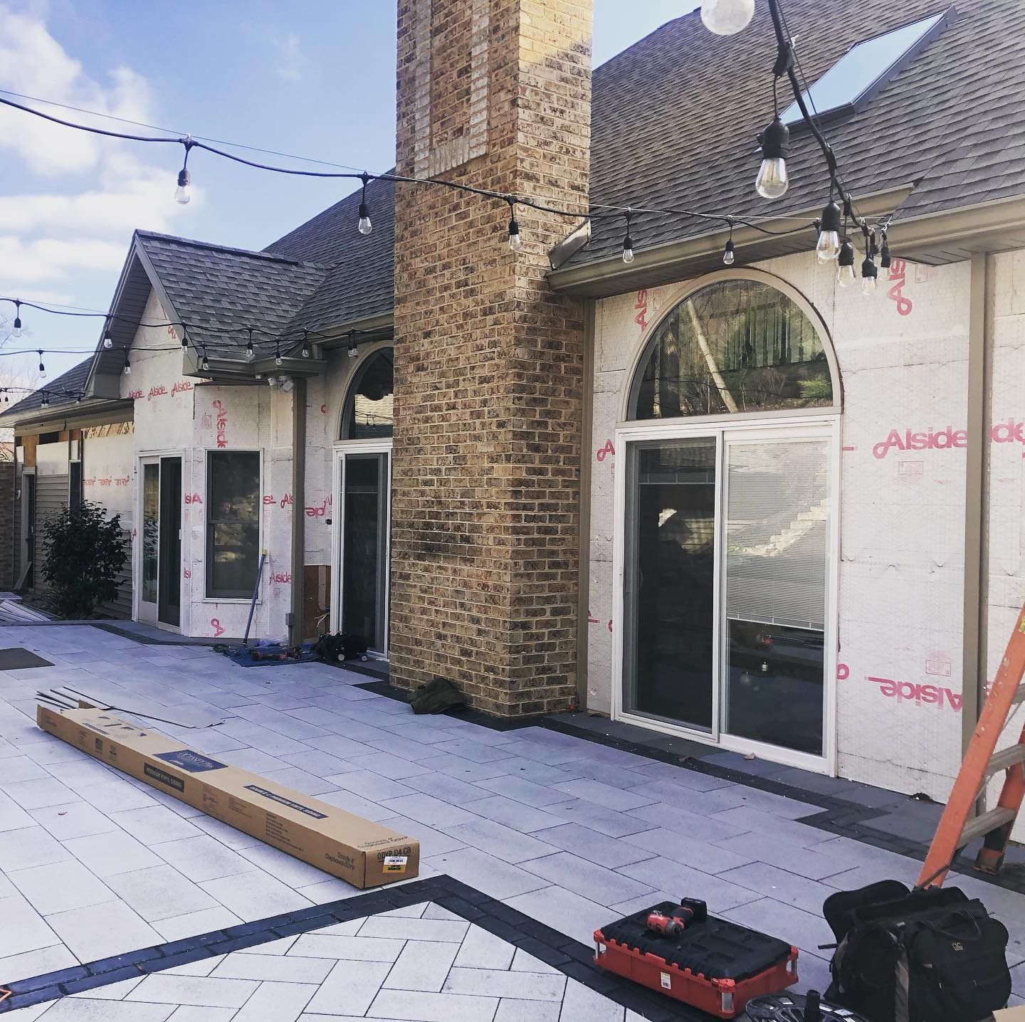 Exterior view of a home under construction with a brick chimney, newly installed patio pavers, and string lights overhead.